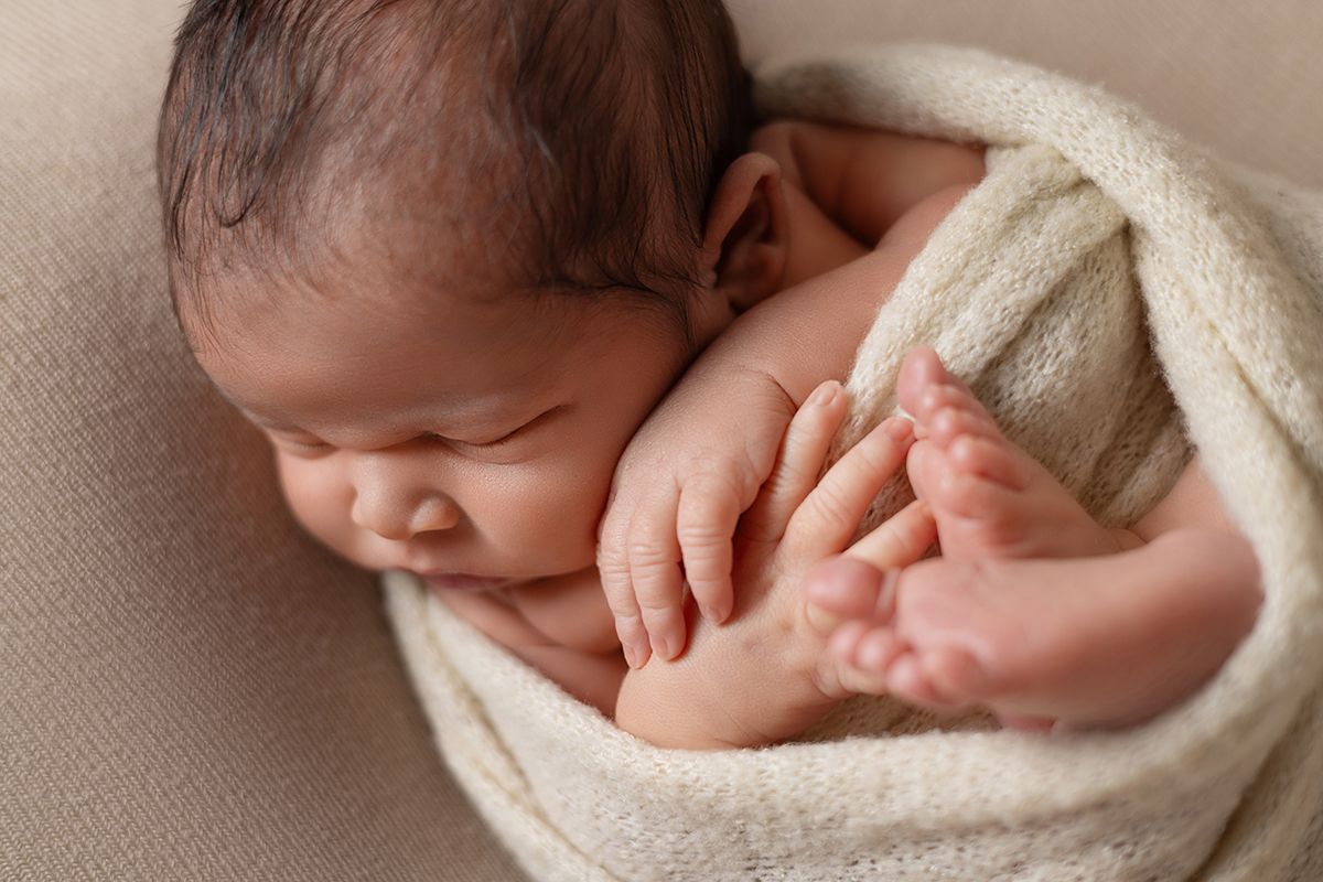 Peaceful newborn baby wrapped in a soft cream blanket, sleeping curled up with tiny hands and feet visible.