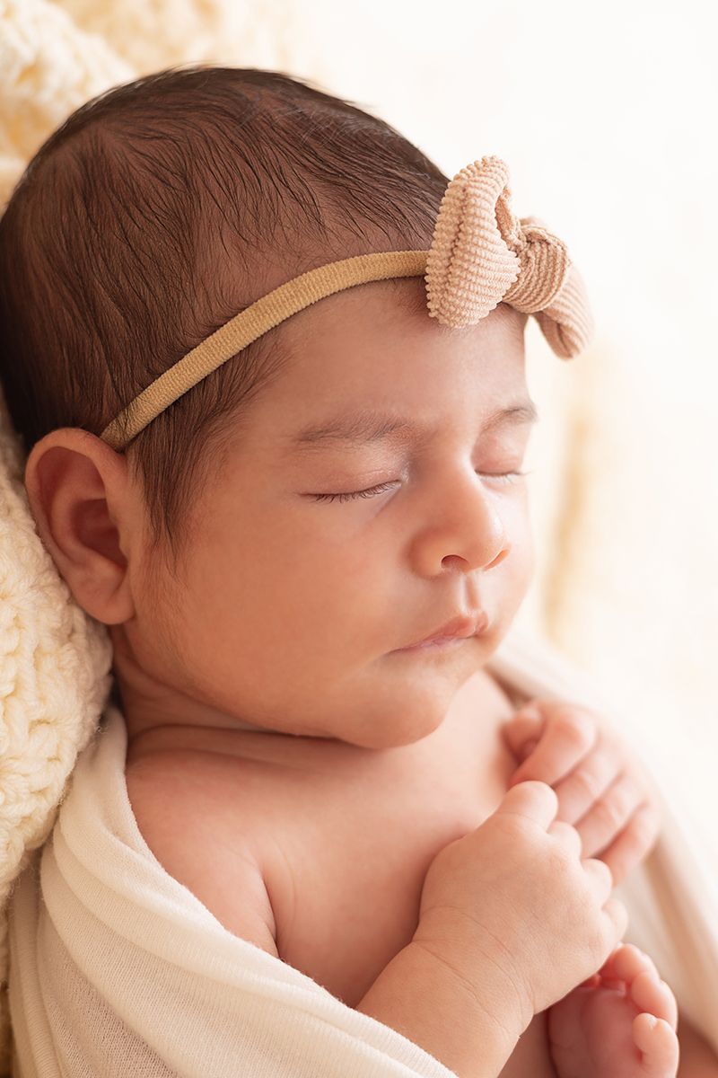 Peaceful sleeping newborn baby girl wearing a beige bow headband, wrapped in a soft cream swaddle blanket.
