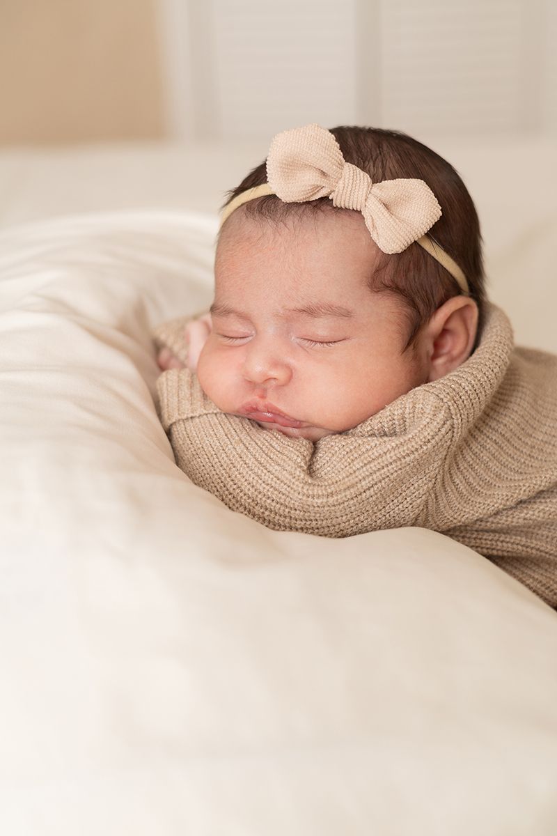 Peaceful newborn baby girl swaddled in a tan knit wrap with a bow headband, sleeping softly.