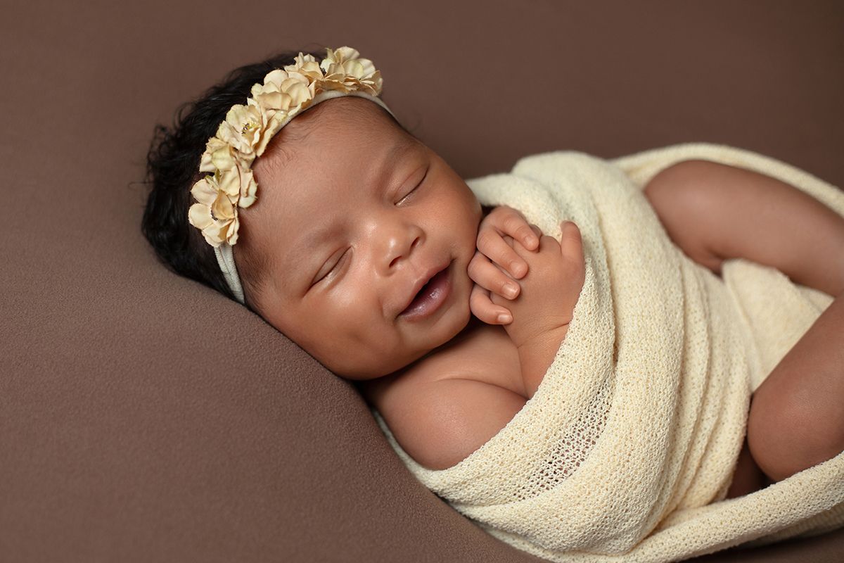 Smiling newborn baby girl wrapped in cream swaddle with floral headband sleeping on brown background.