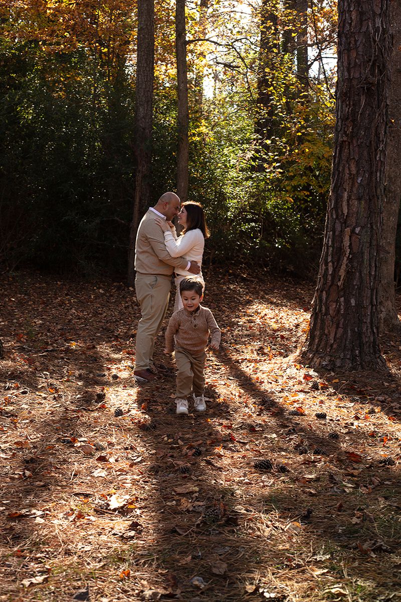A family of three poses in an autumn forest, parents sharing a kiss while their toddler stands in front on a leaf-covered path.