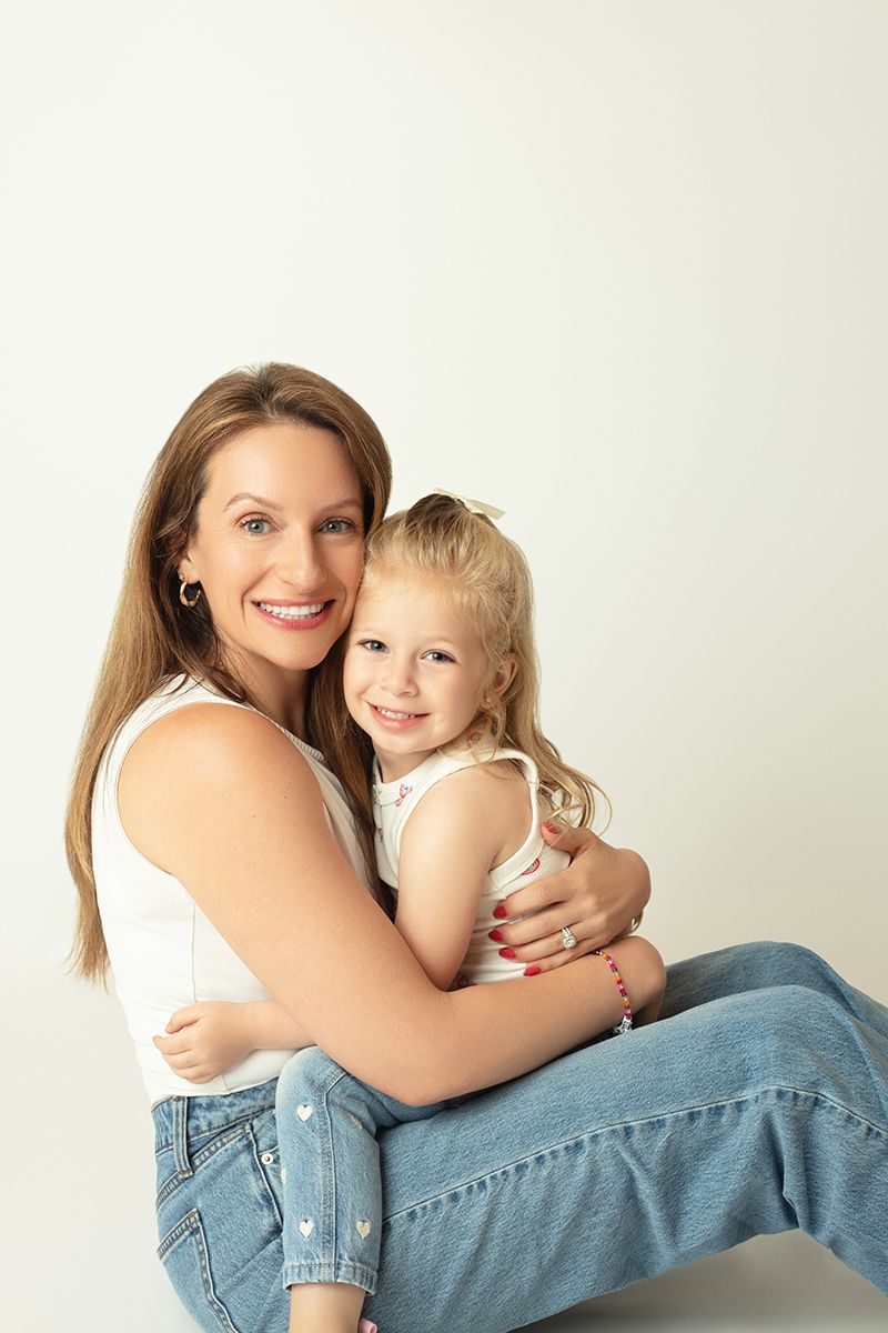 A smiling woman in a white tank top and jeans hugs a young blonde girl in a bright studio setting.