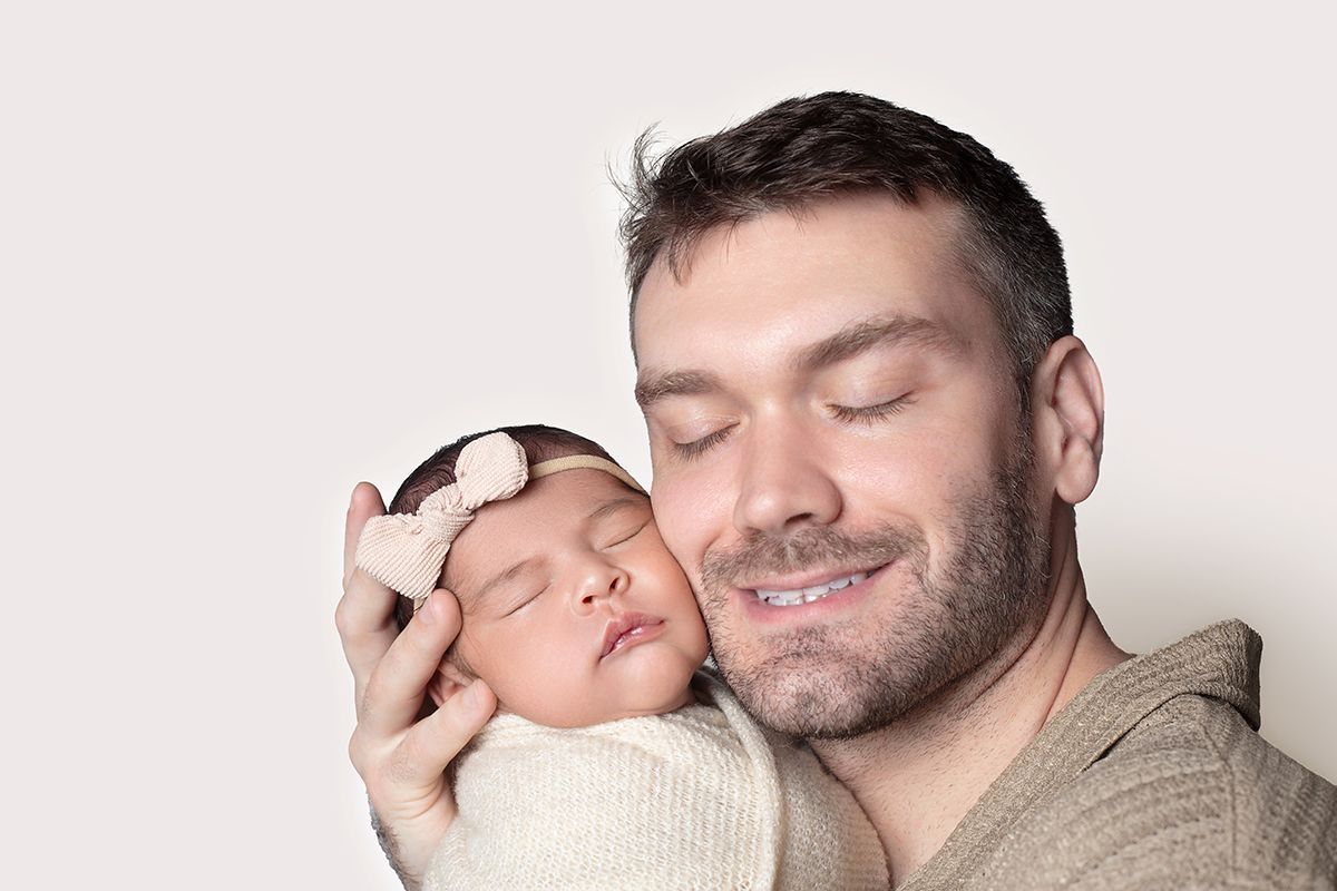 A smiling father with closed eyes gently holds his sleeping newborn baby girl wearing a floral headband.