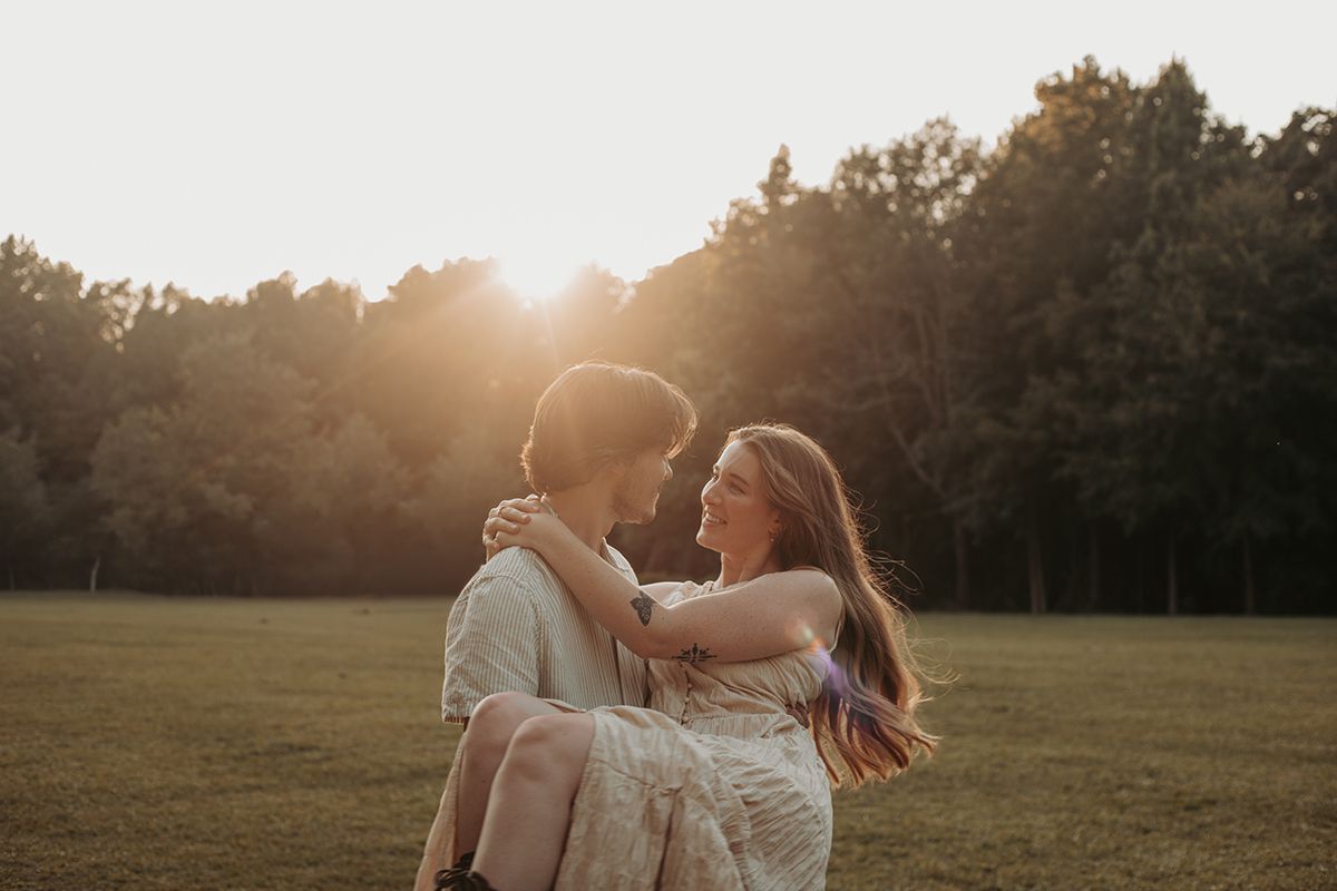 A couple shares a romantic moment at golden hour in a sunlit field, the man lifting the woman as they smile at each other.