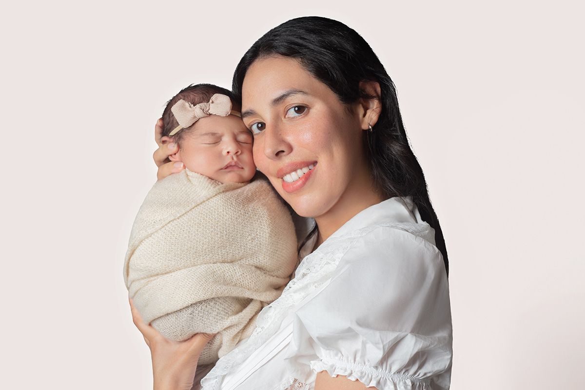 A smiling woman in white holds a swaddled newborn baby wearing a pink bow headband against a soft background.