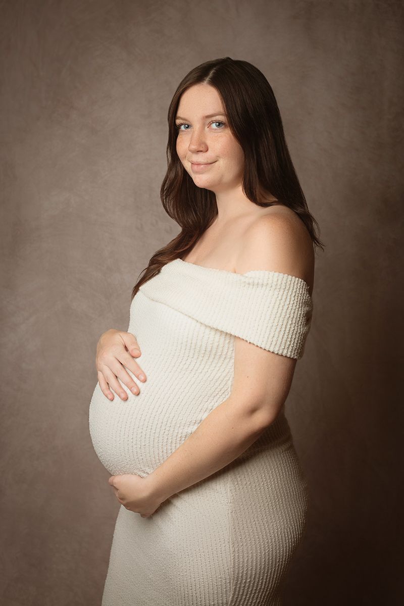 Pregnant woman in white off-shoulder knit dress cradling baby bump, smiling softly against brown studio backdrop.