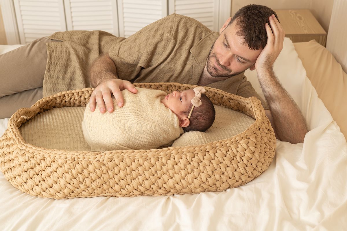Tired father lying on bed watching his swaddled newborn baby sleeping in a woven Moses basket.