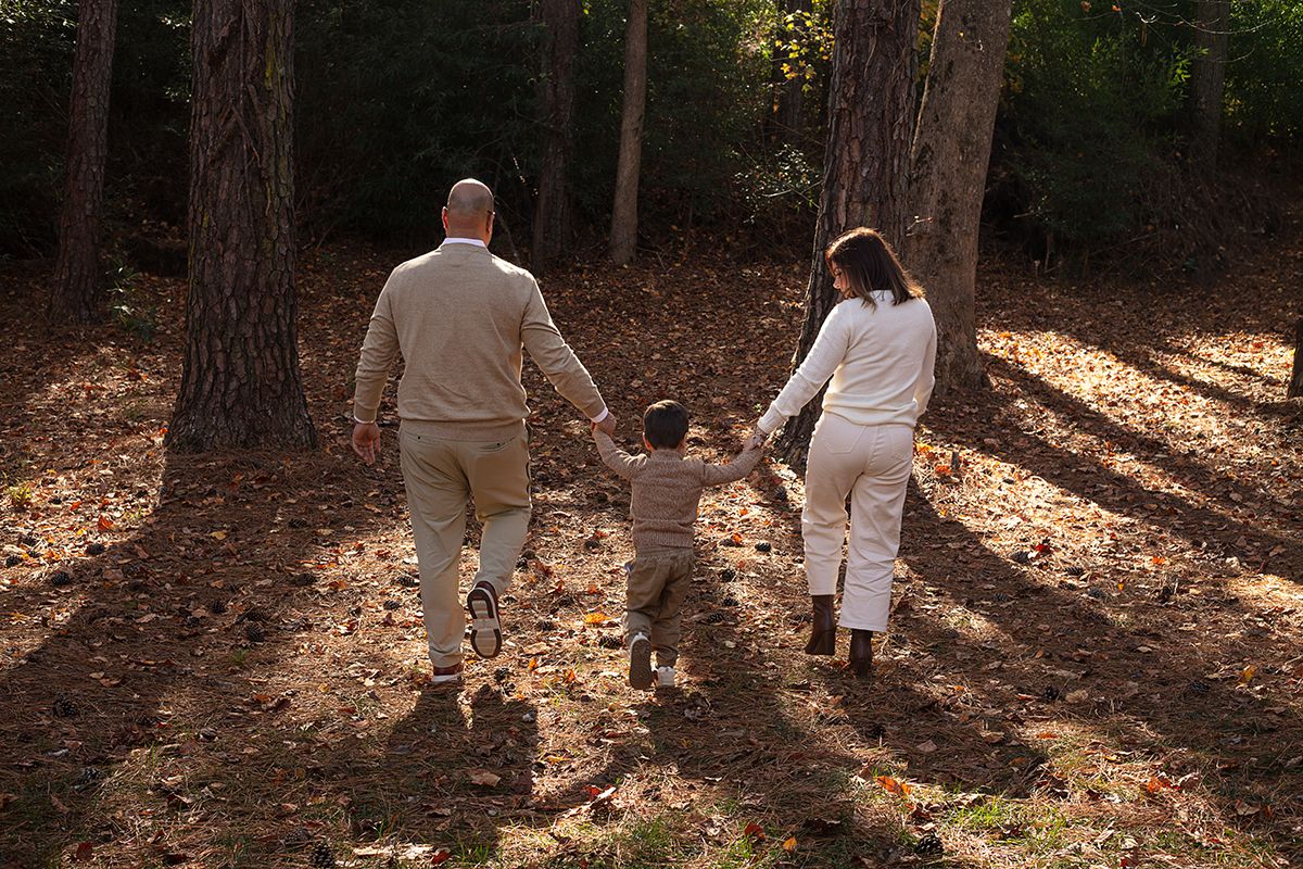 Family of three holding hands walking through a sunlit forest with autumn leaves on the ground.