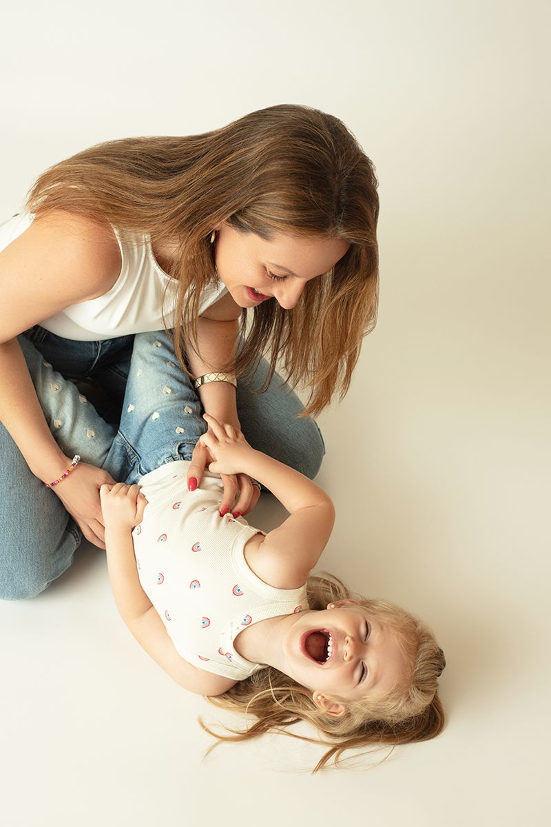 A young mother playfully tickles her laughing toddler daughter on a white studio floor.