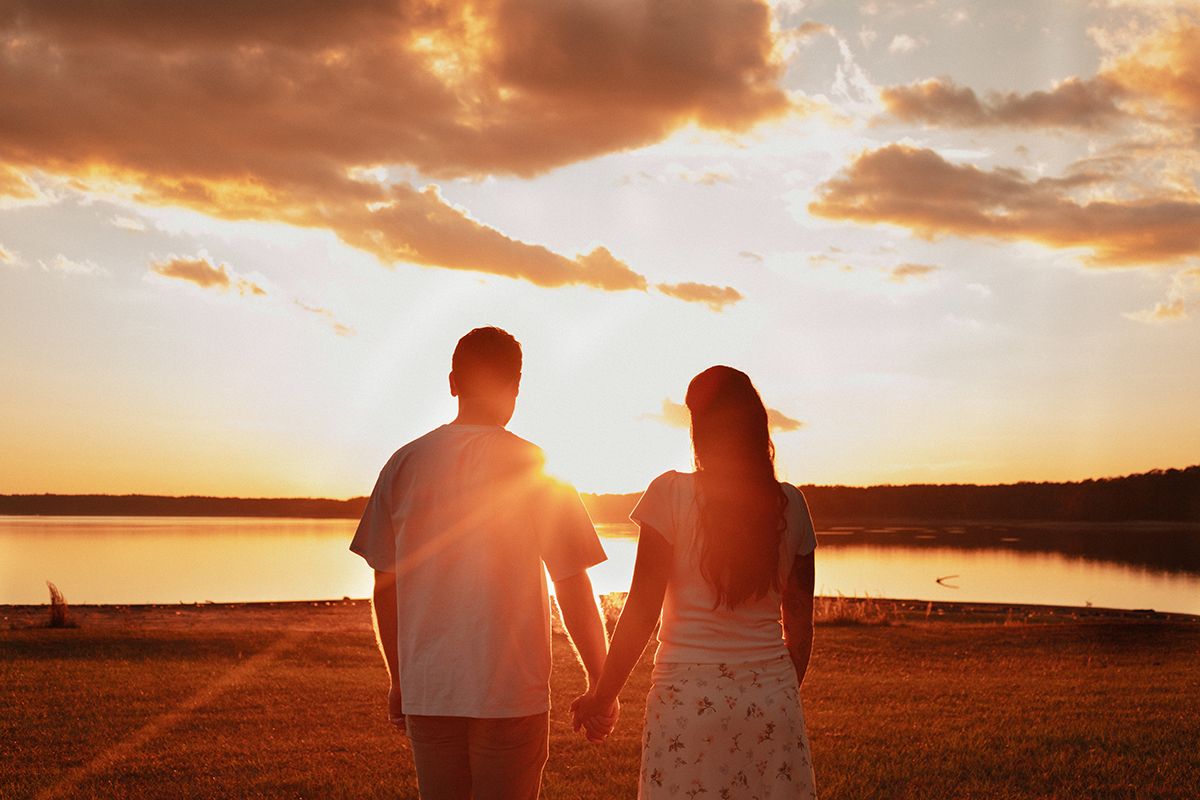 Couple holding hands watching a golden sunset over a calm lake with dramatic clouds in the background.