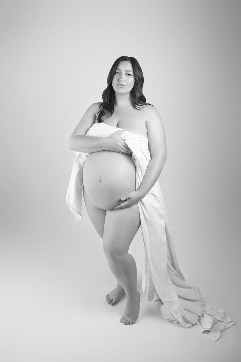 Black and white maternity photo of a pregnant woman draped in white fabric, showcasing her baby bump in a studio setting.