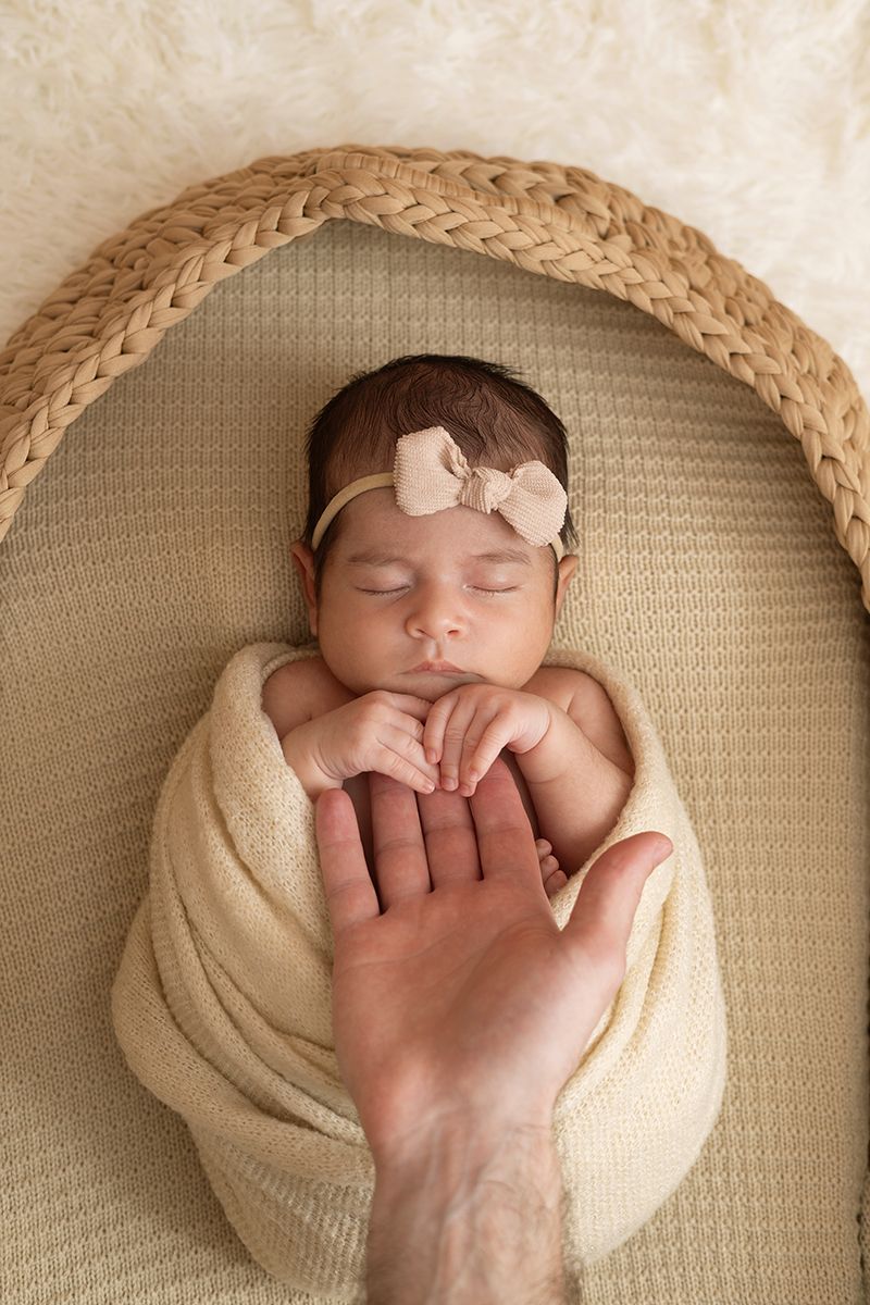 Newborn baby girl sleeping in a woven basket, wrapped in cream, wearing a bow headband, resting on a parent's hand.