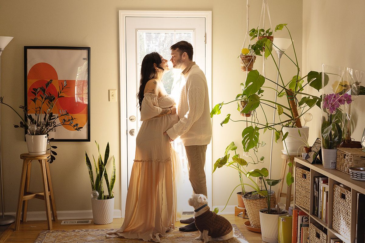 A couple stands close together in a plant-filled home entryway, with a small dog nearby, suggesting a maternity moment.