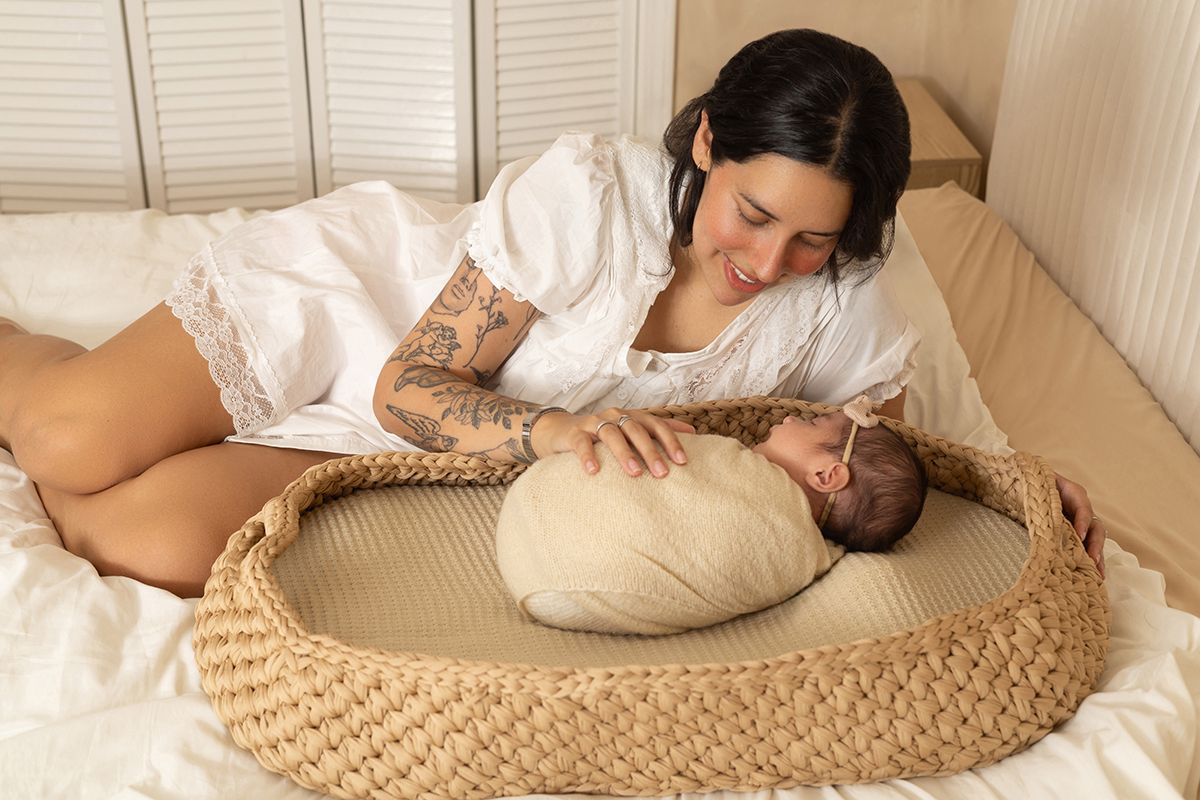 A tattooed mother in white dress gazes lovingly at her swaddled newborn resting in a woven Moses basket on a bed.