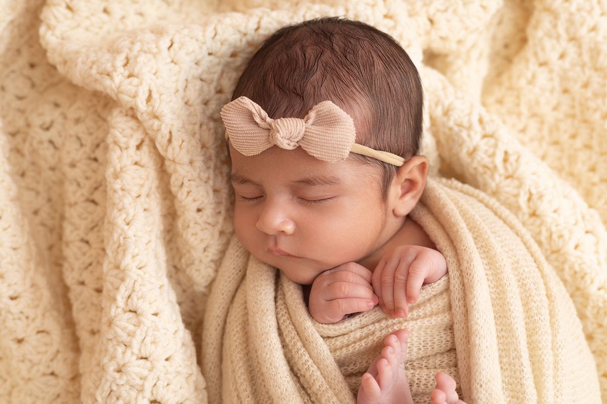 Sleeping newborn baby girl wrapped in cream knit blanket wearing a beige bow headband.