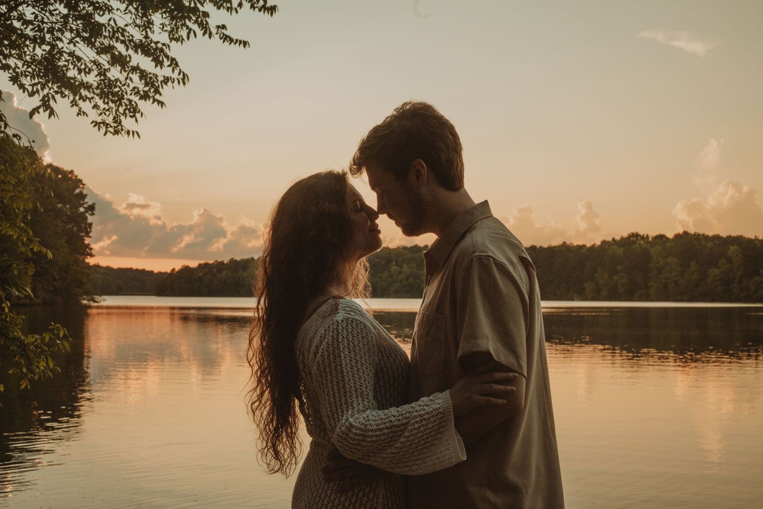 Couple sharing a romantic moment by a serene lake at golden sunset, surrounded by lush trees.