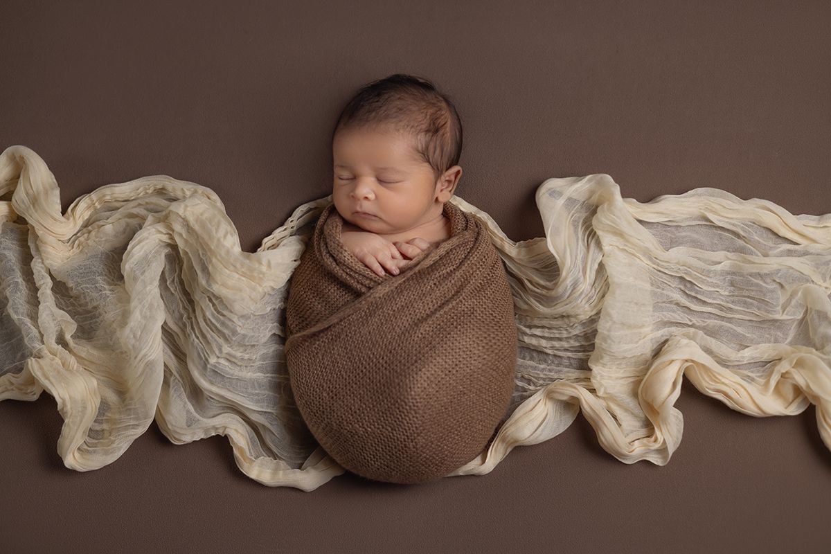 Newborn baby swaddled in brown wrap, sleeping peacefully on a brown backdrop with cream textured fabric.