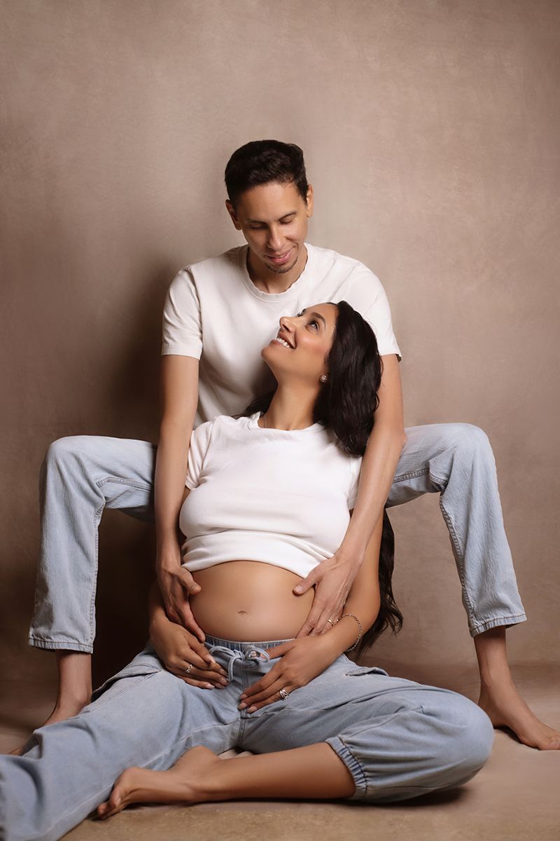 A couple in white tees and jeans pose together, the pregnant woman sitting cross-legged while her partner sits behind her smiling.