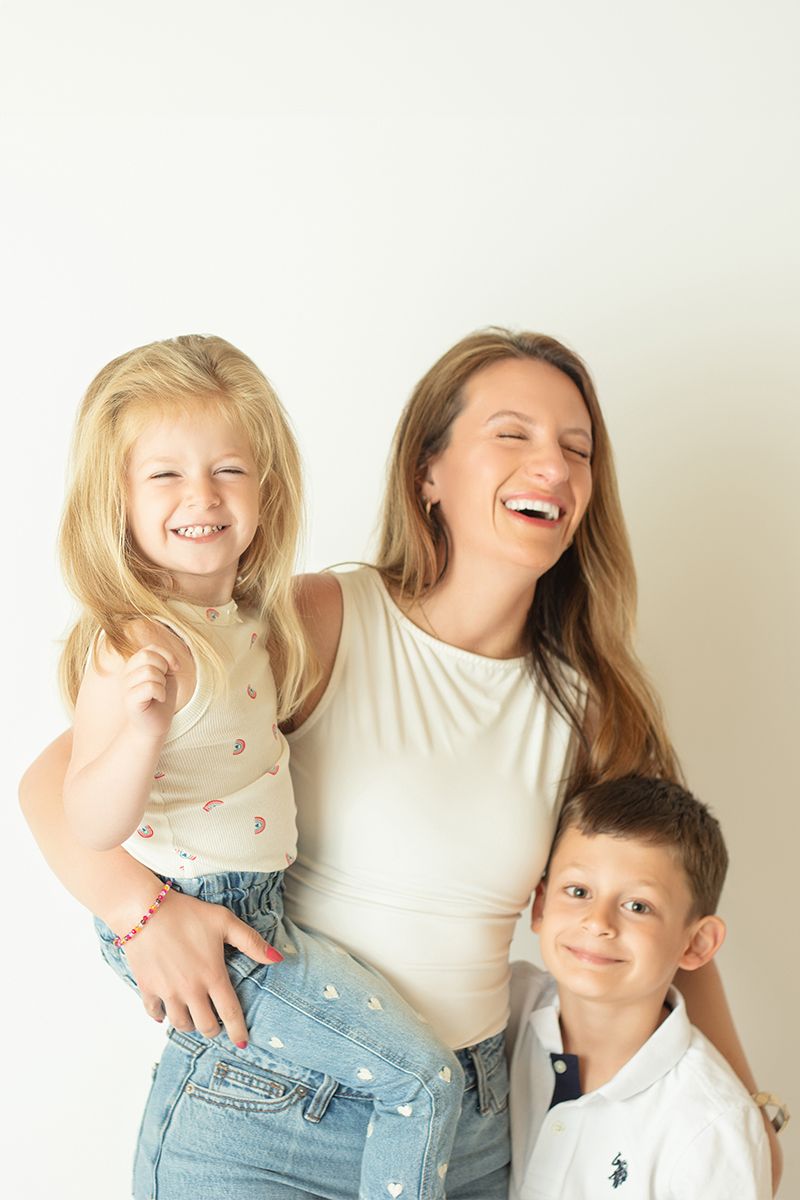 A laughing woman holds a blonde girl while a boy smiles beside them against a white background.