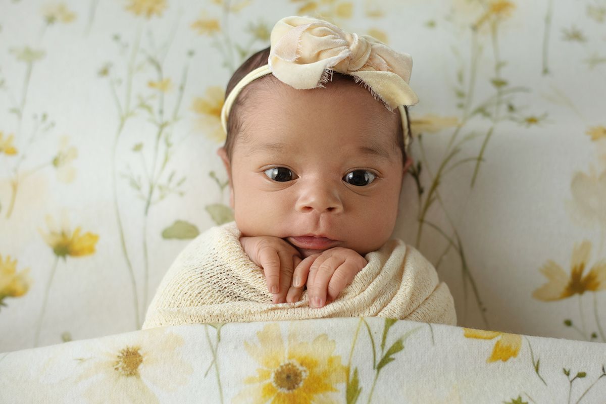 Newborn baby wrapped in cream knit swaddle with floral headband, posed on yellow daisy background.