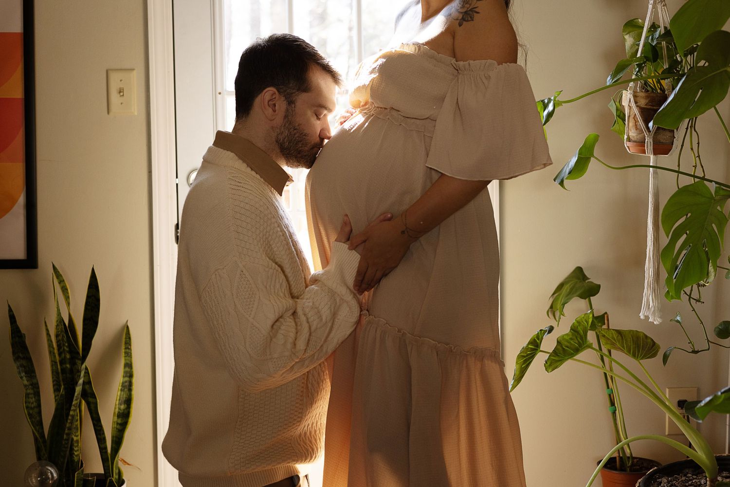 Man kneeling to kiss pregnant partners baby bump in a sunlit doorway surrounded by green houseplants.