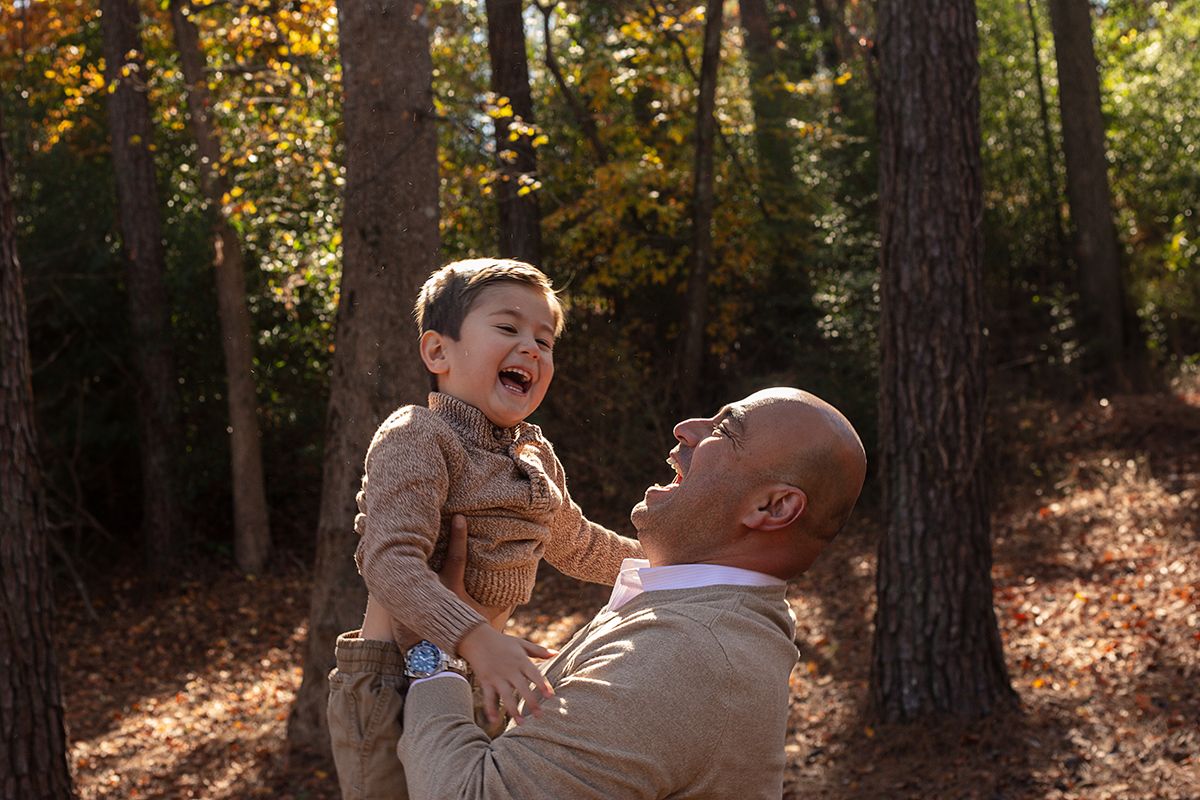 A bald father joyfully lifts his laughing young son among autumn trees in a warm outdoor family moment.