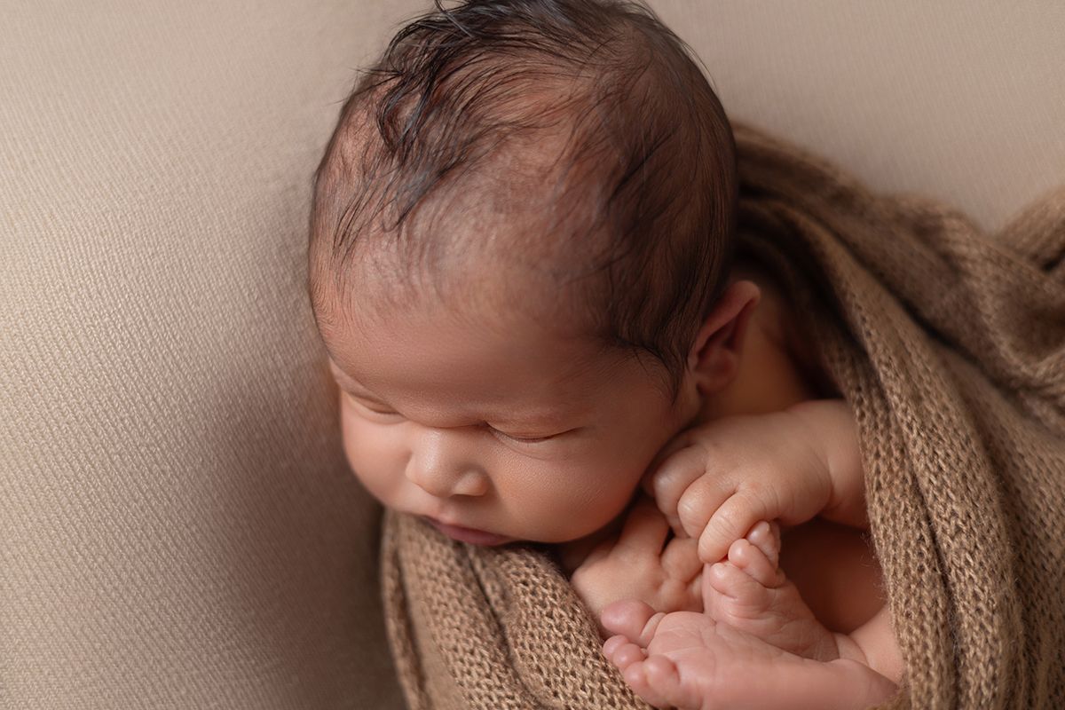Peaceful newborn baby wrapped in a soft brown knit blanket, sleeping with tiny hands visible in a professional photo.