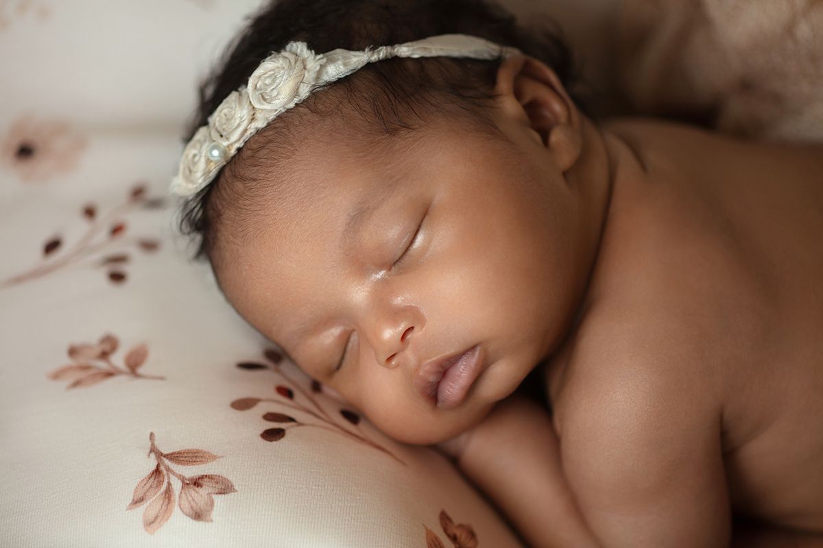 Sleeping newborn baby girl wearing a white floral headband, resting peacefully on a floral patterned blanket.