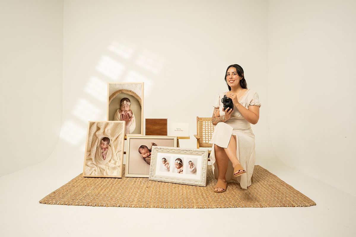Woman sitting beside framed newborn photos and canvas prints displayed on a jute rug in a bright white studio.