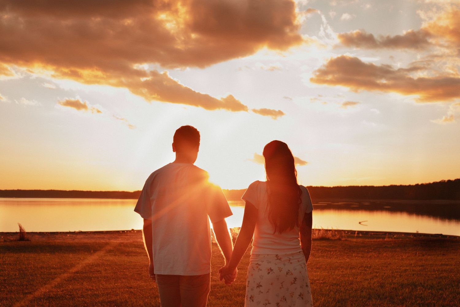 Couple holding hands watching golden sunset over calm lake with dramatic cloudy sky.