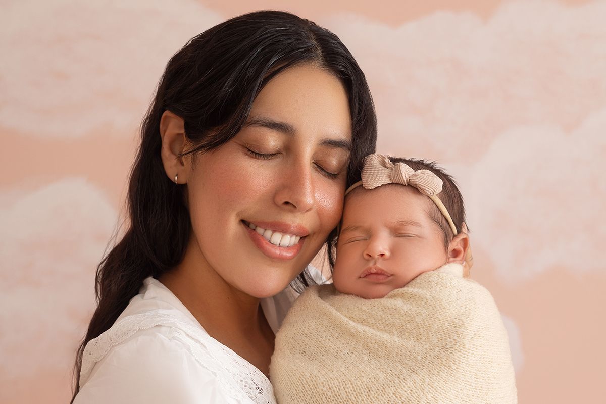 A smiling mother with dark hair holds her sleeping newborn baby wrapped in a cream swaddle with a pink bow headband.