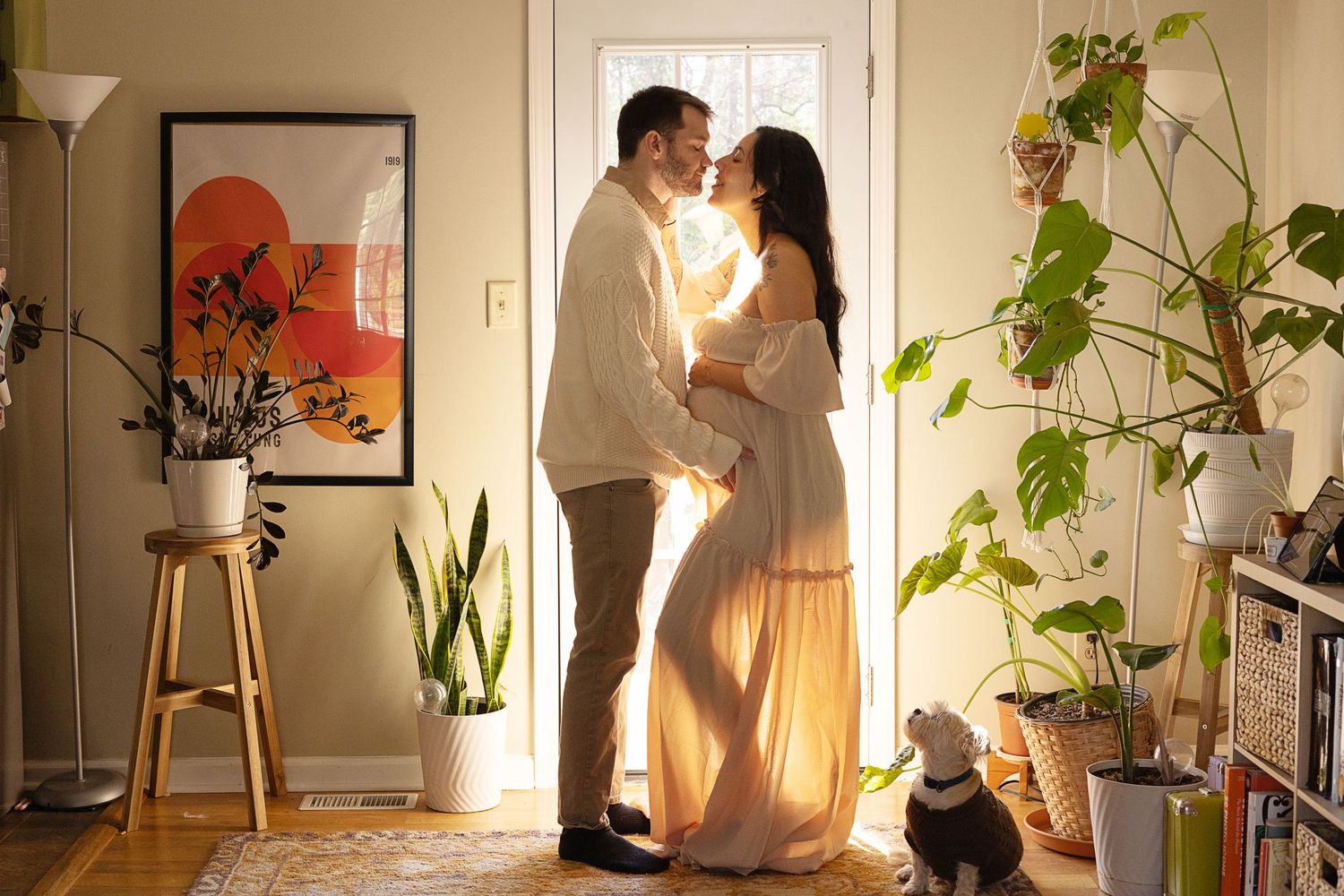 Couple sharing a tender moment in a sunlit bohemian living room surrounded by lush plants, with a small dog nearby.