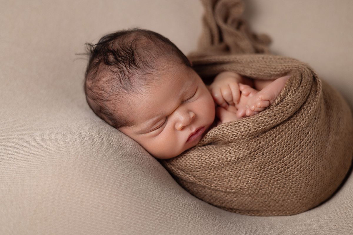 Newborn baby swaddled in brown wrap sleeping peacefully on beige background in professional newborn photography.
