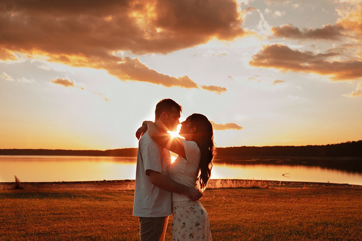 Couple embracing at golden sunset by a lake, silhouetted against vibrant orange sky with dramatic clouds.
