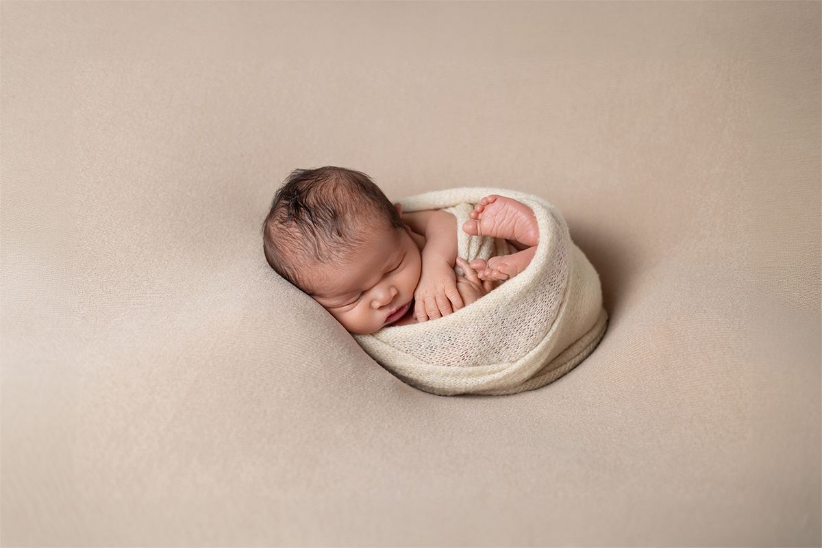 Newborn baby sleeping peacefully curled in a cream wrap bowl prop on a beige backdrop.