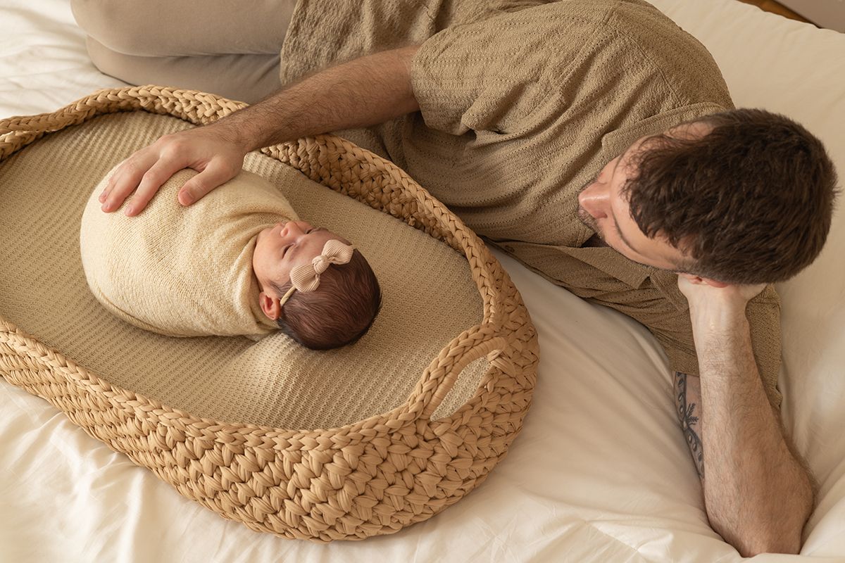 Father gazing lovingly at swaddled newborn baby resting in a woven Moses basket on a white bed.