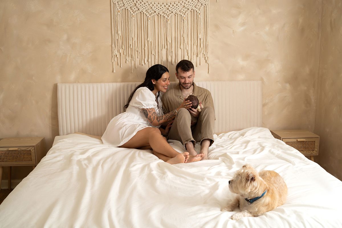 Couple sitting on a white bed looking at a phone together with a fluffy dog resting nearby in a cozy bedroom.