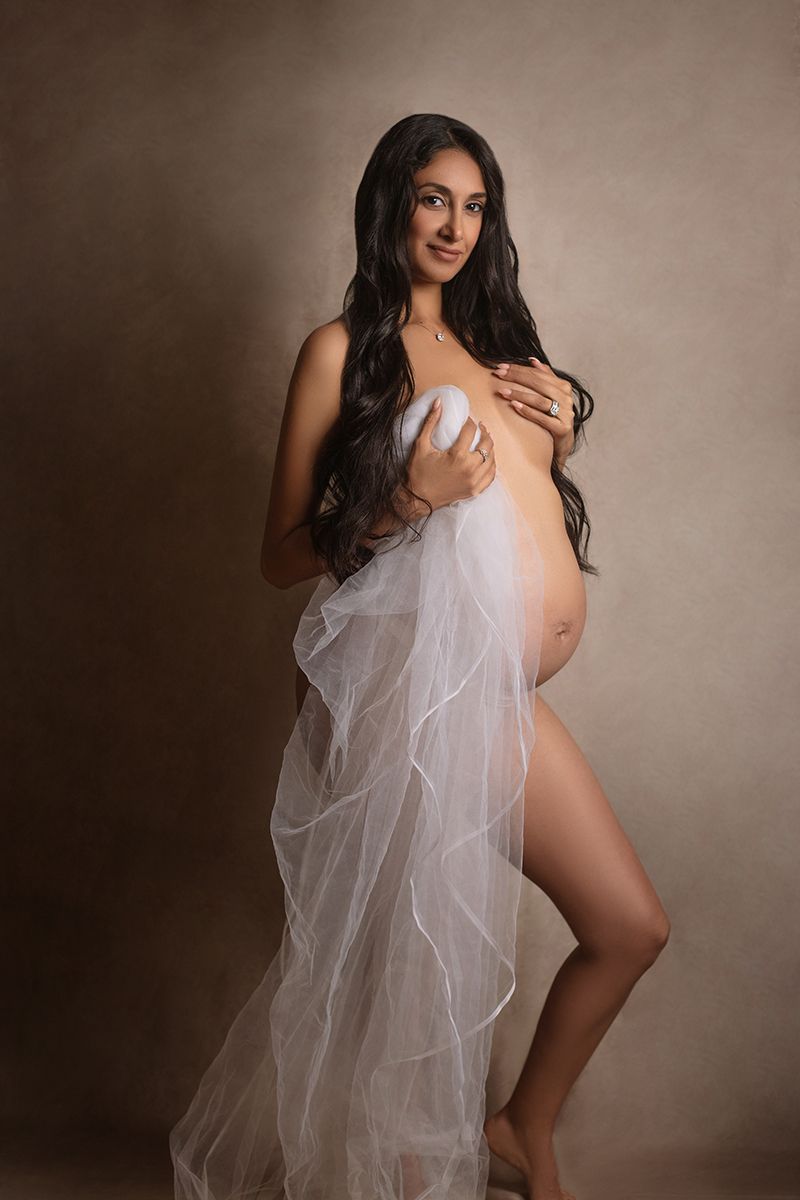 Pregnant woman posing elegantly with white tulle fabric in a professional maternity photography studio session.
