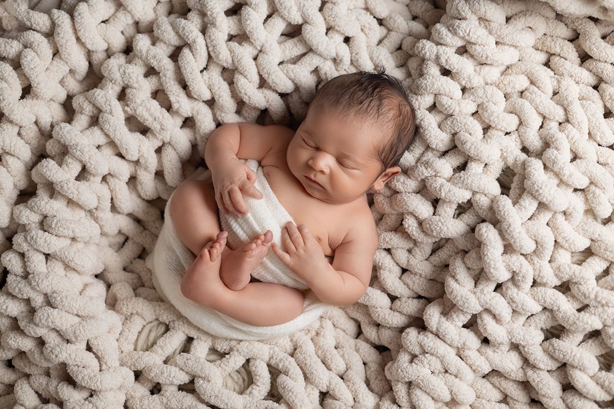 Sleeping newborn baby wrapped in white swaddle holding a milk bottle on a chunky knit cream blanket.