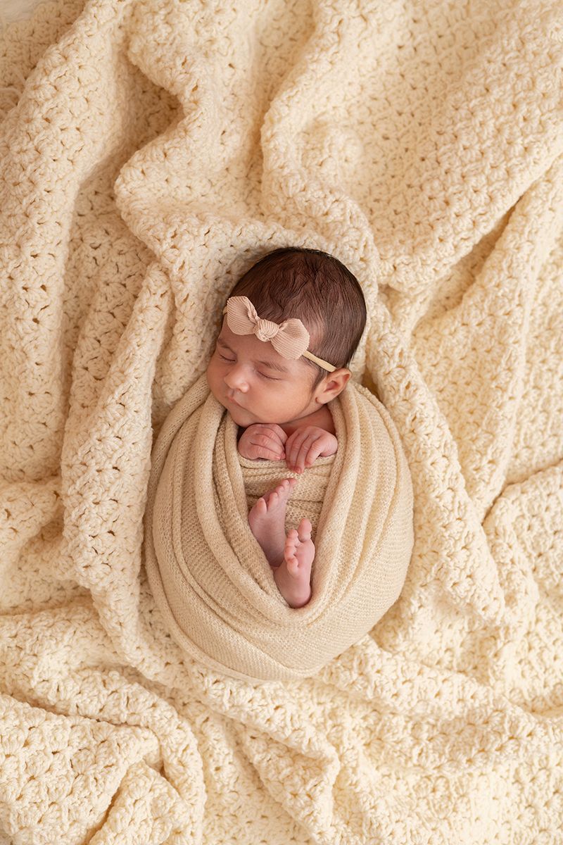 Sleeping newborn baby girl swaddled in a cream wrap with a bow headband on a soft knit blanket.