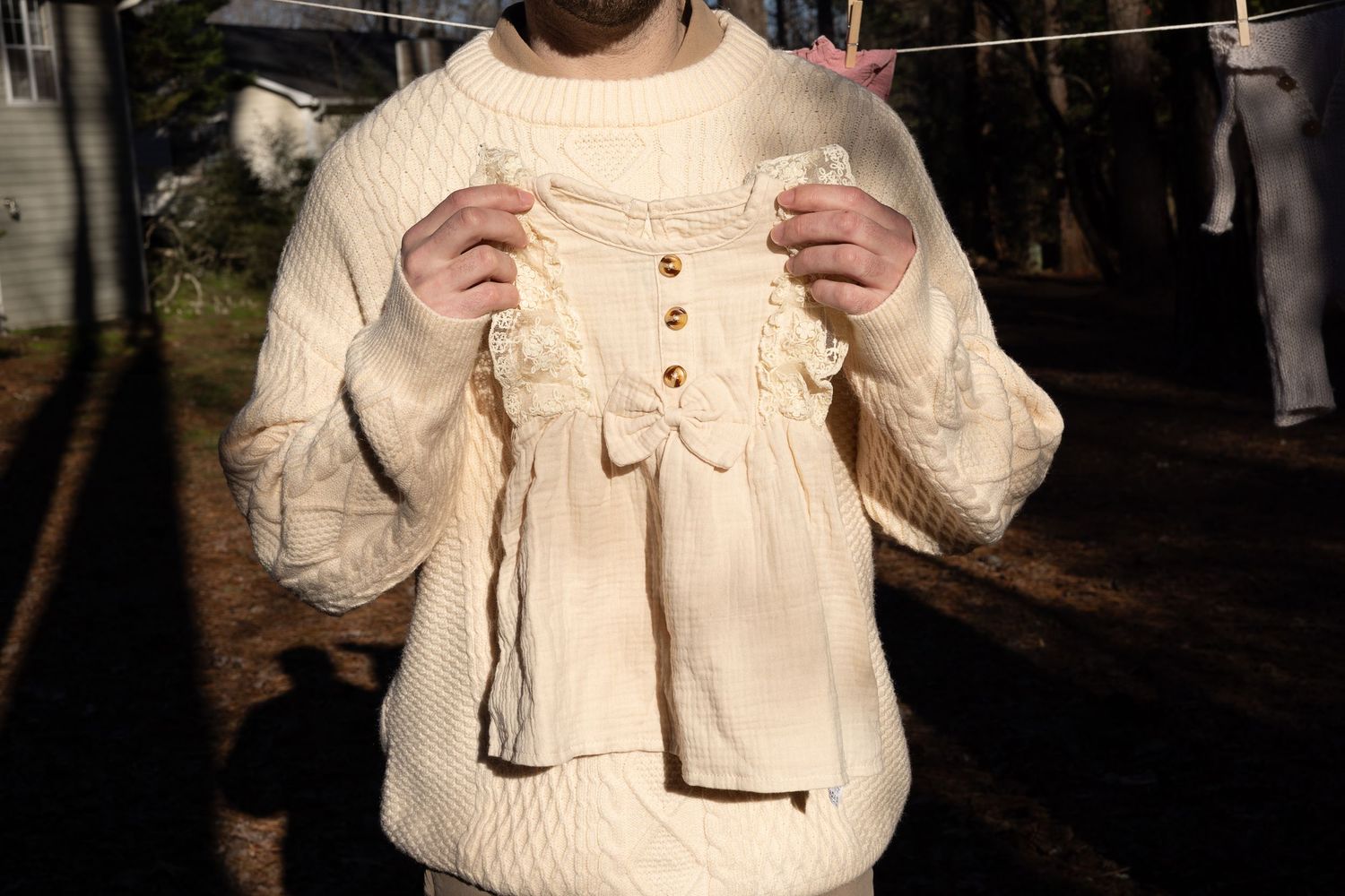 Person in cream knit sweater holding up a delicate ivory baby dress with buttons and bow outdoors.