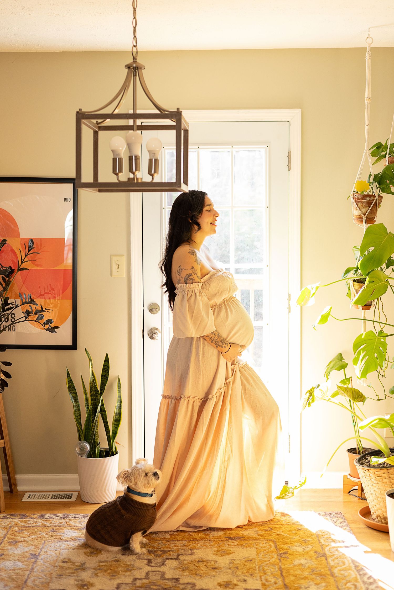 Pregnant woman in an off-shoulder peach gown standing by a sunlit door with her dog nearby.