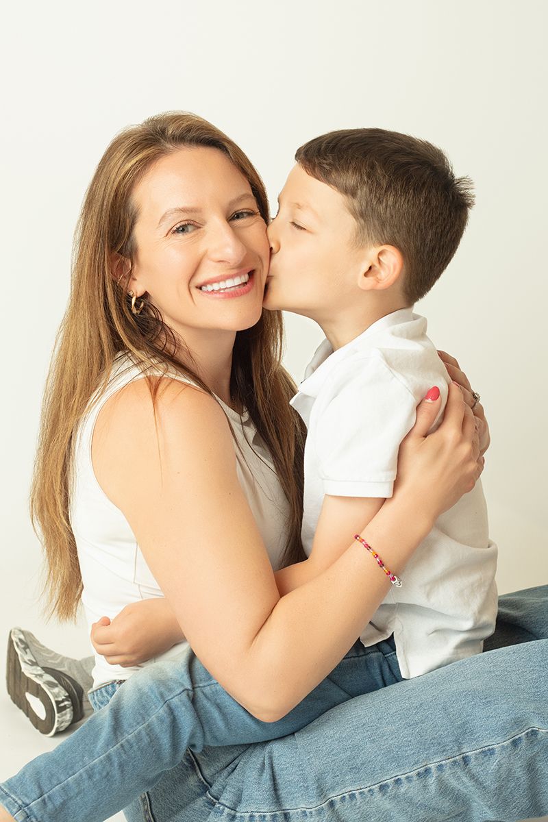 A smiling woman in a white top receives a kiss on the cheek from a young boy in a white shirt.