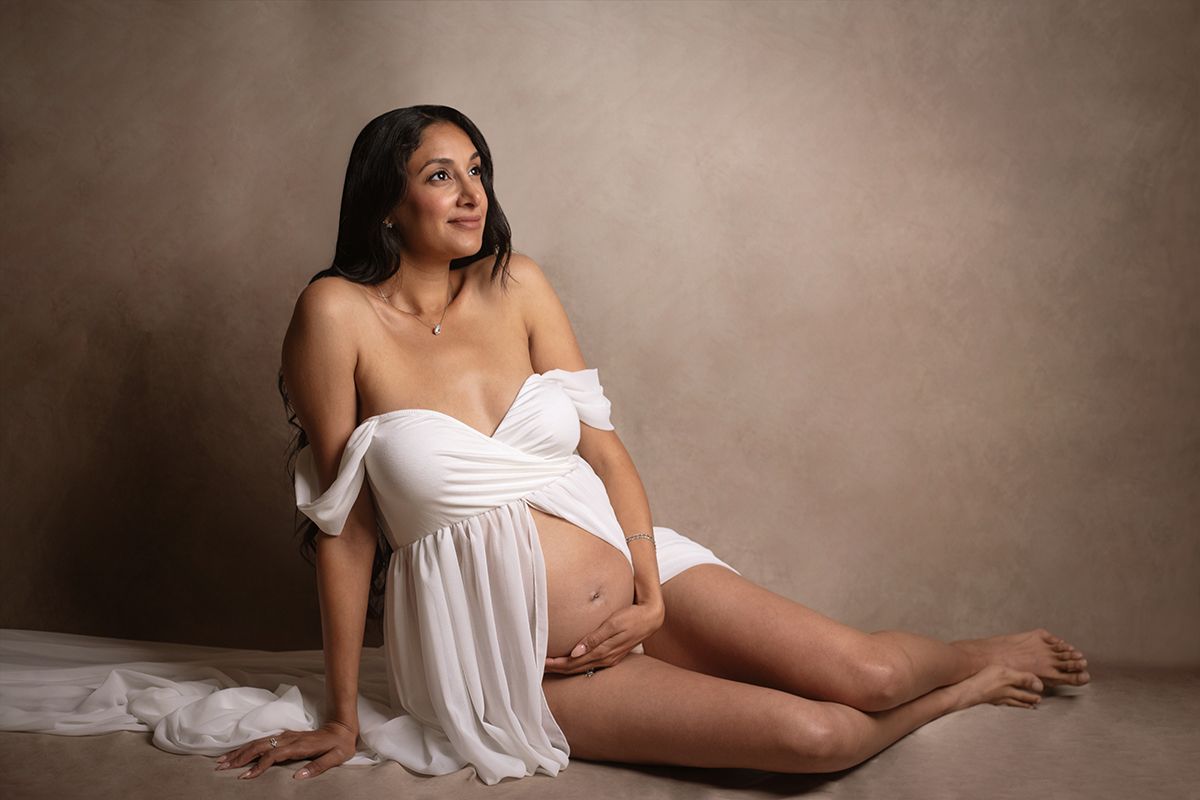Pregnant woman in white draped fabric poses gracefully on studio floor against neutral backdrop.