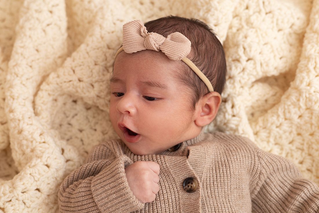 Newborn baby girl in beige knit outfit and bow headband yawning on cream blanket.