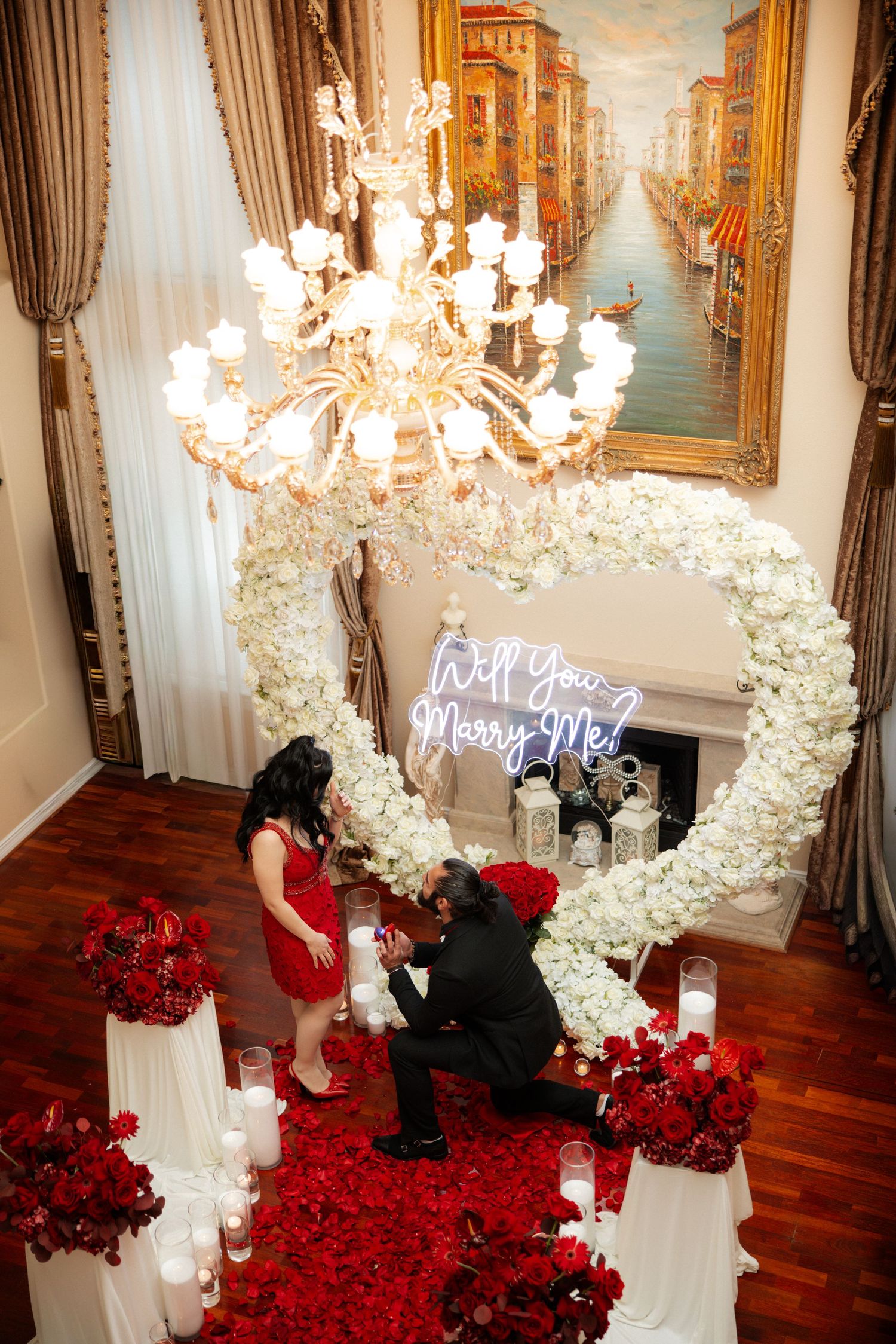 A romantic proposal setup with red roses and white floral arch under a crystal chandelier.