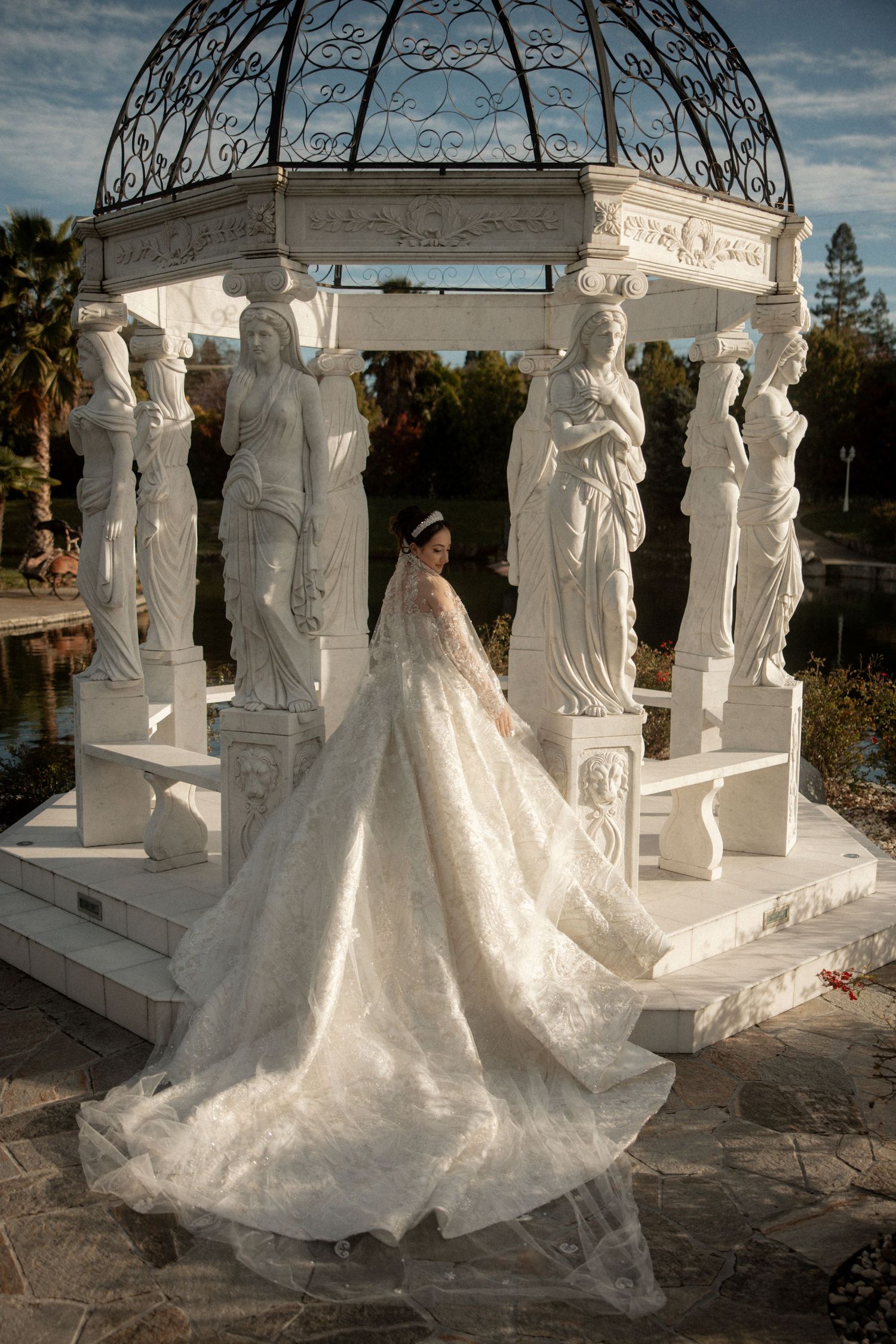 Bride in ornate lace wedding gown with cathedral train posing at classical white gazebo with statues.