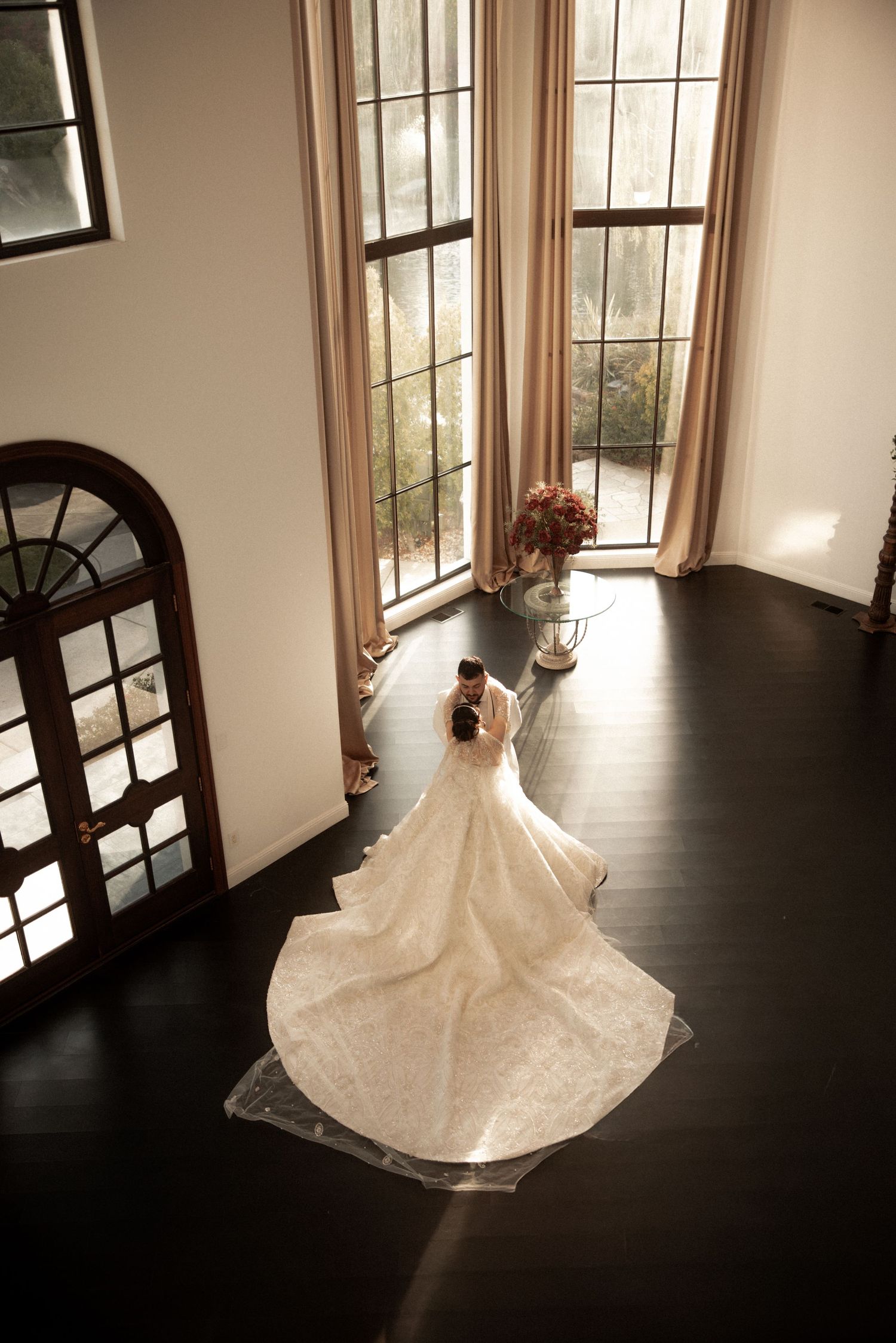 Aerial view of bride in long white wedding gown standing near tall windows in elegant venue.