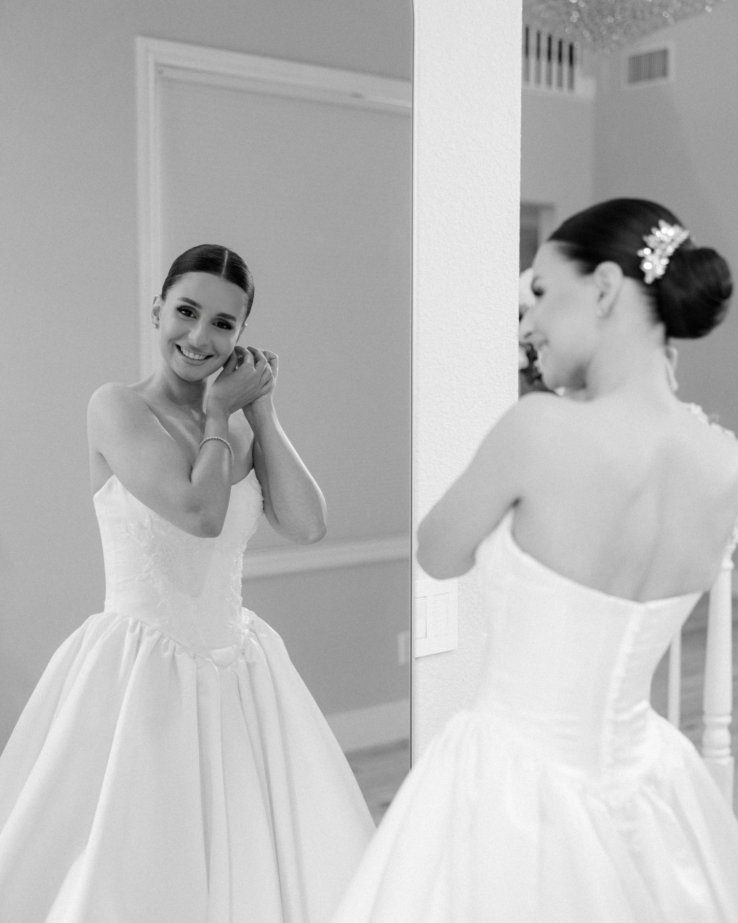 Black and white photo of a smiling bride adjusting her earring in front of a mirror.