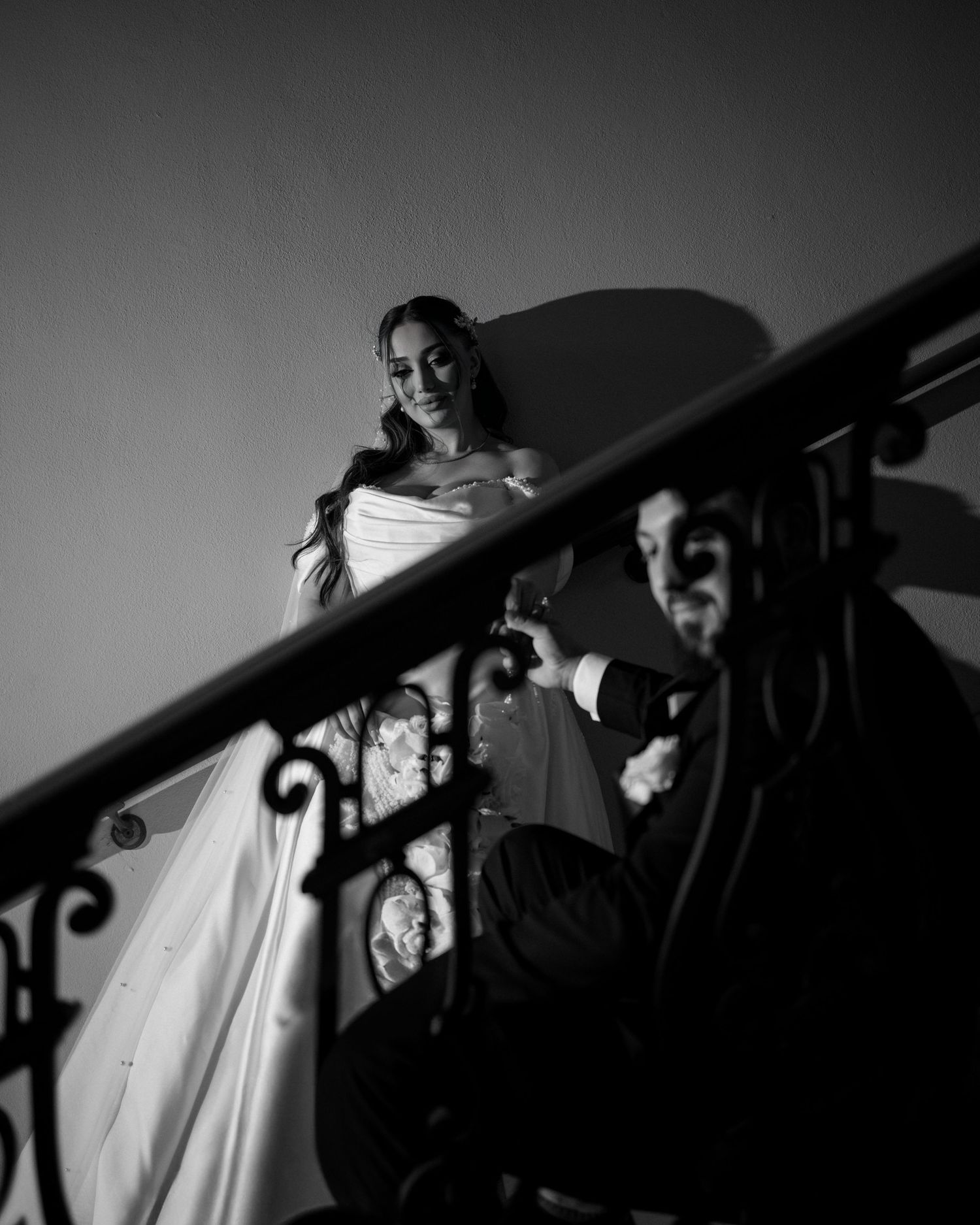 A dramatic black and white photo of a bride descending an ornate staircase with wrought iron railings.